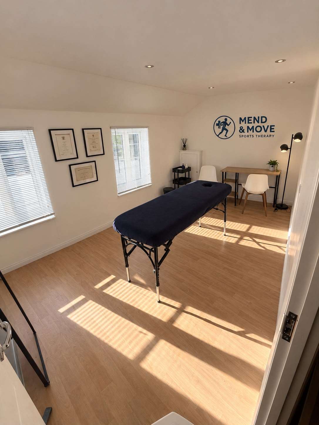 The Mend & Move treatment room in Hythe: a treatment couch in the sunlight, framed qualifications and the Mend & Move logo on the wall